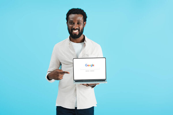 KYIV, UKRAINE - MAY 17, 2019: smiling african american man pointing at laptop with google website, isolated on blue