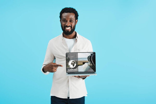 smiling african american man pointing at laptop with booking website isolated on blue