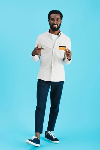 smiling african american man pointing at credit card isolated on blue