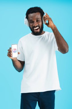 Kiev, Ukrayna - 17 Mayıs 2019: gülümseyen Afro-Amerikan adam kulaklıkile müzik dinliyor ve apple music uygulaması ile akıllı telefon gösteren, mavi izole