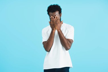 frustrated african american man in white t-shirt closing face, isolated on blue   clipart