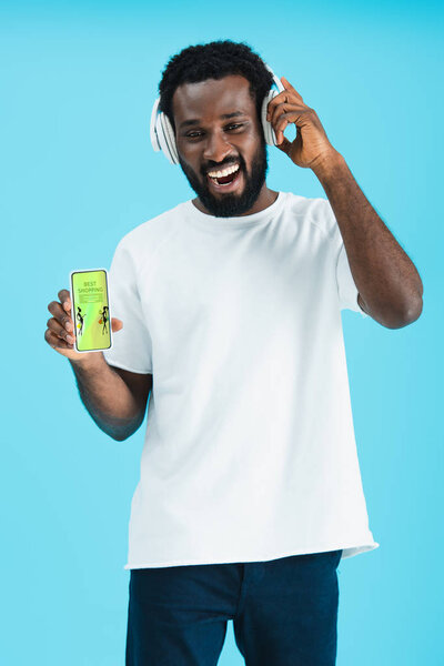 smiling african american man listening music with headphones and showing smartphone with best shopping app, isolated on blue