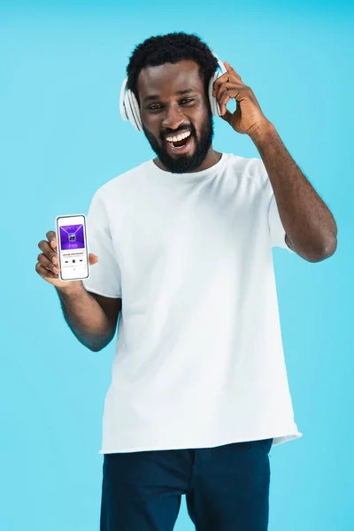 乌克兰 2019年5月17日 微笑的非洲裔美国男子听音乐与耳机和显示智能手机与苹果音乐应用程序 隔离在蓝色 — 图库照片