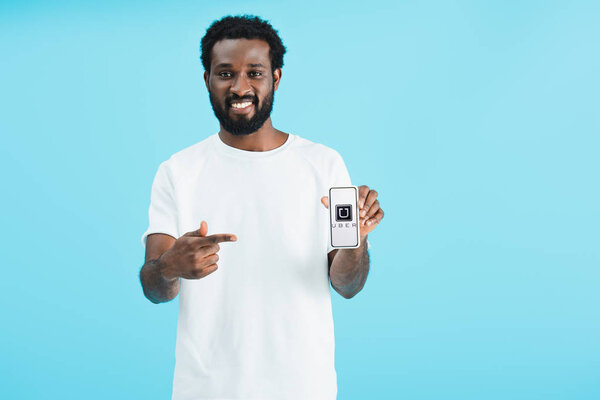 KYIV, UKRAINE - MAY 17, 2019: smiling african american man pointing at smartphone with uber app, isolated on blue