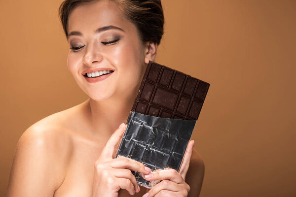 beautiful smiling young naked woman holding chocolate bar in silver foil isolated on beige