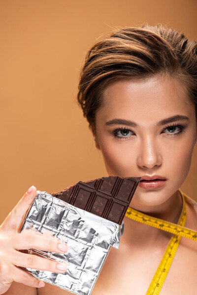 naked young woman with yellow measuring tape holding chocolate bar isolated on beige