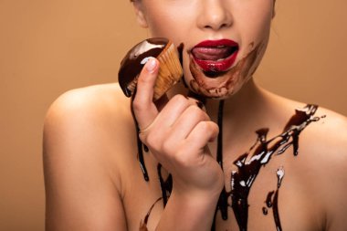 partial view of naked woman licking red lips with chocolate spills on skin and holding muffin isolated on beige clipart