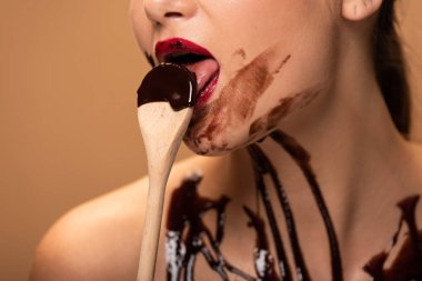 cropped view of naked woman with red lips and chocolate spills on skin licking spoon isolated on beige clipart