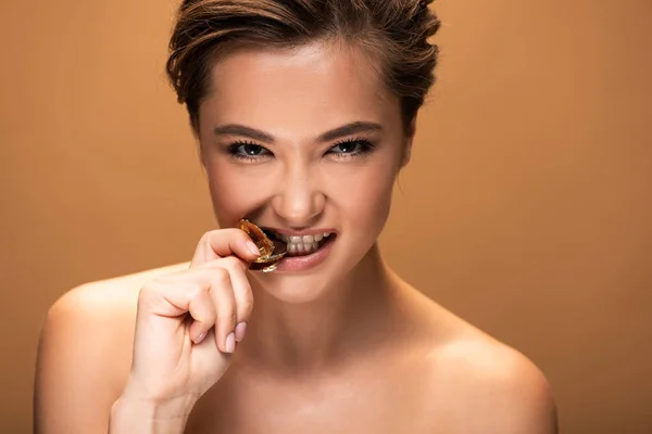 ベージュに隔離された金色の箔でチョコレートコインを噛む裸の若い女性 — ストック写真