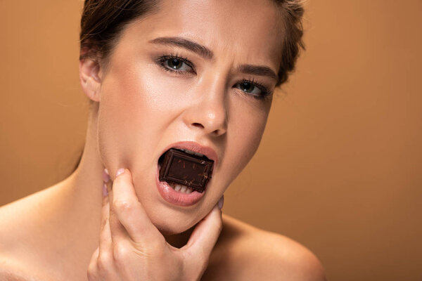 stresses woman with chocolate piece in mouth holding face with hand isolated on beige 