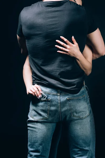 back view of woman hugging man in jeans and taking out condom from pocket isolated on black