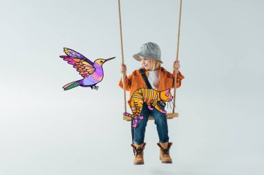 cute kid in jeans and orange shirt sitting on swing with fantasy cat on knees and looking at colorful bird illustration on grey background clipart