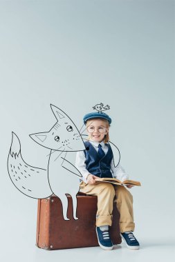 smiling kid in retro vest with fantasy bird on cap sitting on suitcase near fairy fox and holding book on grey background clipart