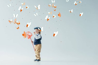 cute and blonde kid in retro vest and cap holding butterfly net on grey background with fairy butterflies clipart