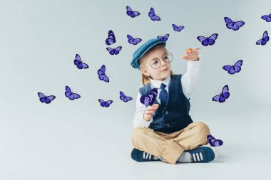 gri arka plan üzerinde peri mor kelebekler arasında çapraz bacaklar ile katta oturan sevimli çocuk 