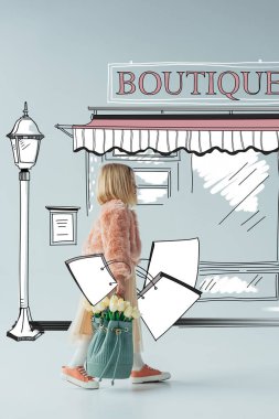 side view of cute kid with white tulips and shopping bags looking at fairy boutique on street on grey background clipart