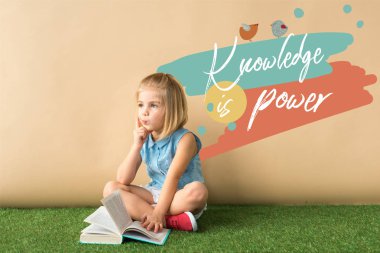 thoughtful cute child sitting with crossed legs on grass rug and holding book on beige background with knowledge is power lettering  clipart