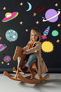 happy child in trench coat and jeans sitting on rocking horse on black background with fairy cosmic illustration clipart