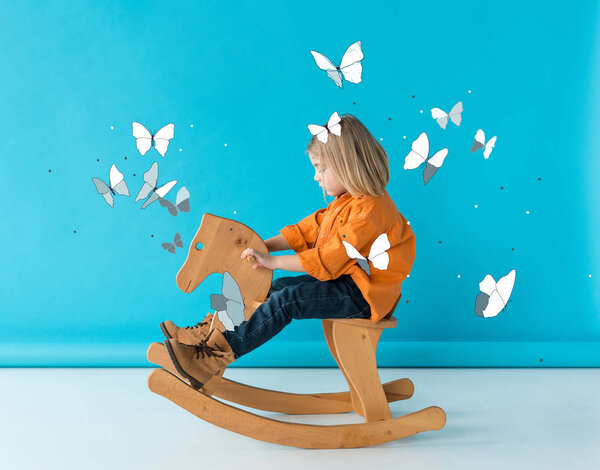 kid in jeans and orange shirt riding rocking horse on blue background with fairy butterflies illustration