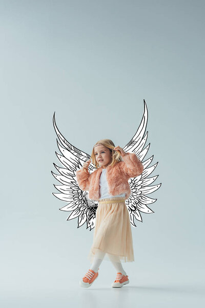 cute kid in faux fur coat and skirt with fairy angel wings walking and looking away on grey background