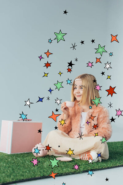 smiling child sitting with crossed legs on grass rug, playing with toy dishes among stars illustration isolated on grey