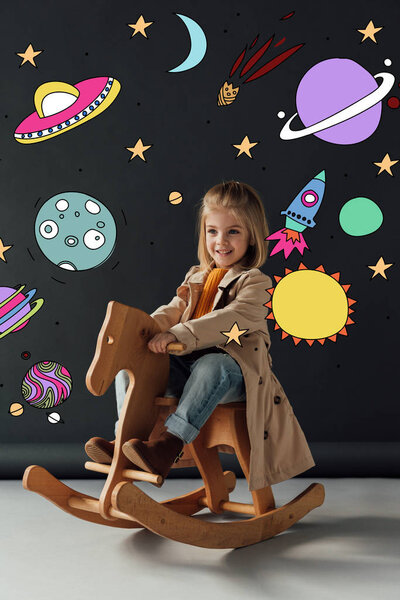 happy child in trench coat and jeans sitting on rocking horse on black background with fairy cosmic illustration