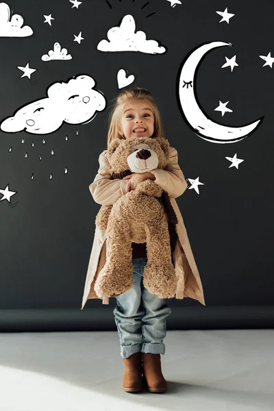 トレンチコートとジーンズで幸せでかわいい子供は魔法の月 星と雨の雲のイラストと黒の背景にテディベアを抱きしめる — ストック写真