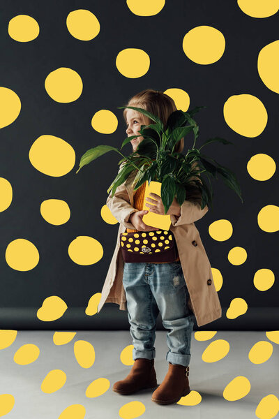  cute child in trench coat and jeans holding plant in flowerpot on black background with yellow abstract dots illustration