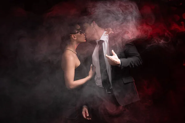 选择性的焦点女孩在礼服和面具接吻与男朋友在烟雾在黑色 — 图库照片