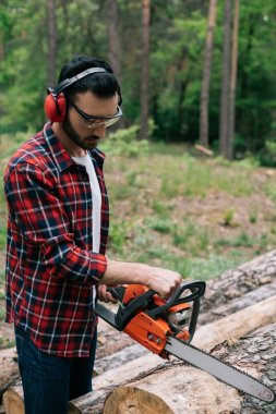 attentive lumberer in checkered shirt cutting wood with chainsaw in forest clipart