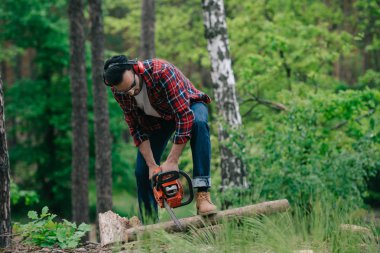 lumberjack in plaid shirt and denim jeans cutting wood with chainsaw in forest clipart