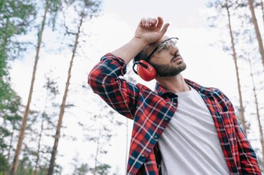 tired lumberjack in protective glasses holding hand near head and looking away in forest clipart