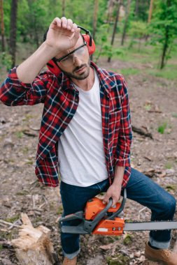 exhausted lumberjack with chainsaw holding hand near head and looking away in forest clipart