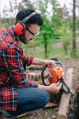 lumberman in noise-canceling headphones fixing chainsaw in forest clipart
