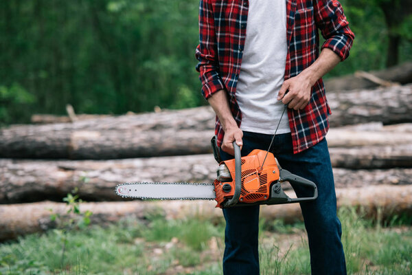 partial view of lumberjack in checkered shirt holding chainsaw in forest