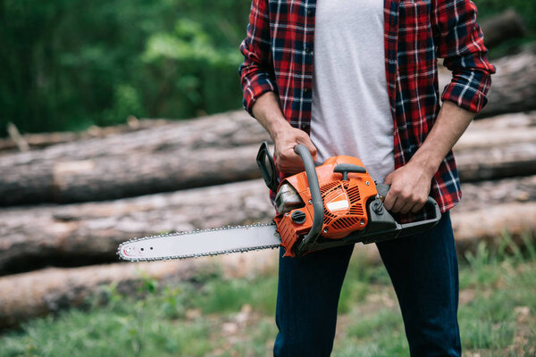 cropped view of lumberjack holding chainsaw while standing near logs in forest