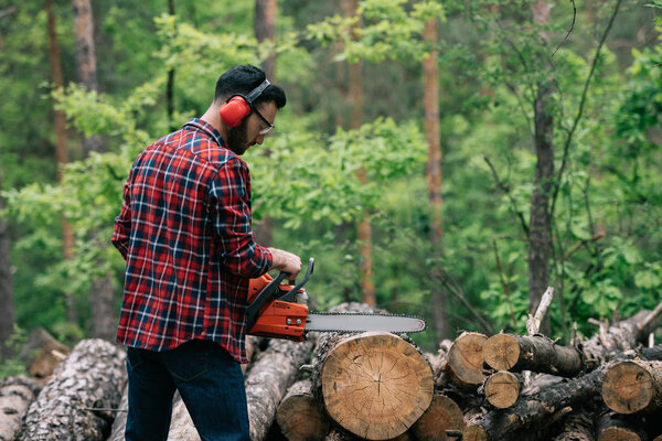 bearded lumberer in plaid shirt cutting trunks with chainsaw in wood