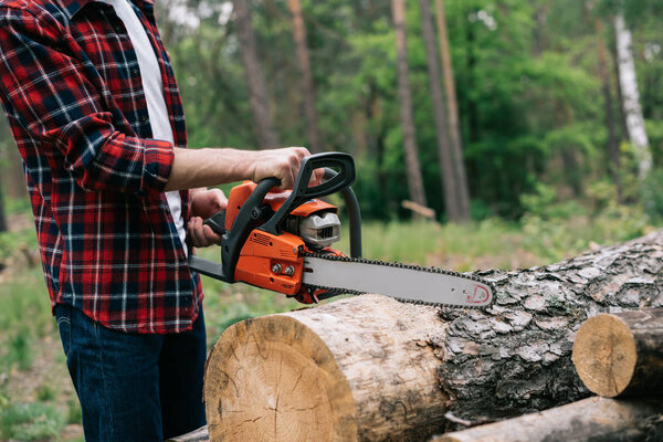 partial view of lumberer in checkered shirt cutting round timber with chainsaw in forest