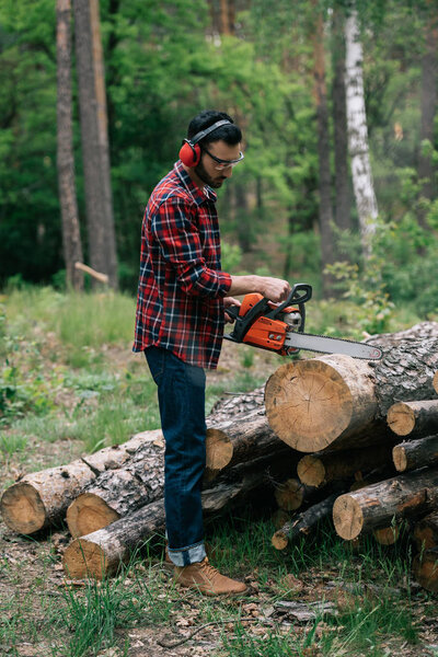 bearded lumberjack in noise-canceling headphones cutting wood with chainsaw in forest