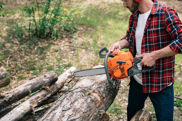 lumberer in plaid shirt cutting round timbers with chainsaw in forest