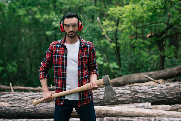 serious lumberjack in protective glasses and noise-canceling headphones holding ax and looking at camera