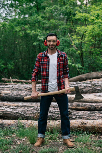 cheerful lumberjack in protective glasses and noise-canceling headphones holding ax and smiling at camera