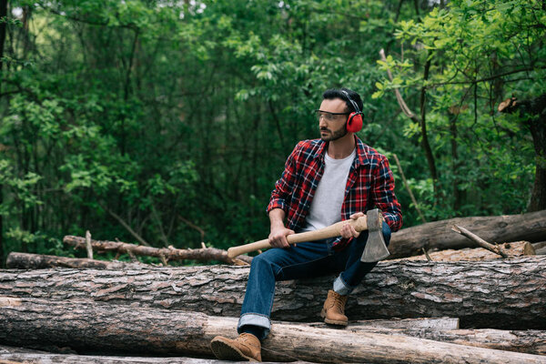 pensive lumberman with ax sitting on logs in forest and looking away