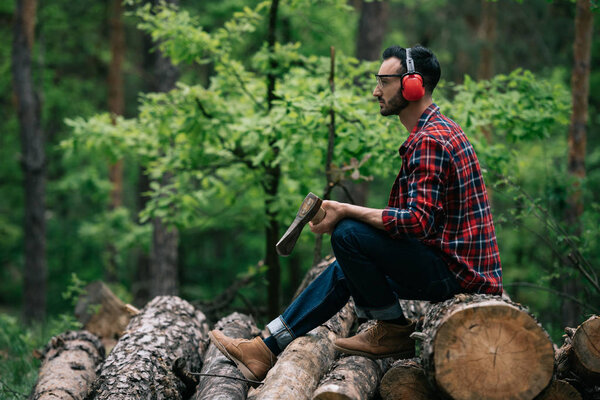 thoughtful lumberman holding ax while sitting on logs in forest and looking away