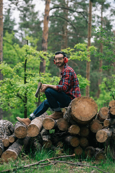 cheerful lumberjack holding ax while sitting on tree trunks in forest and smiling at camera