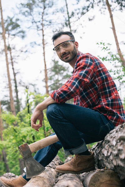 smiling lumberer looking at camera while sitting on logs in forest and holding ax