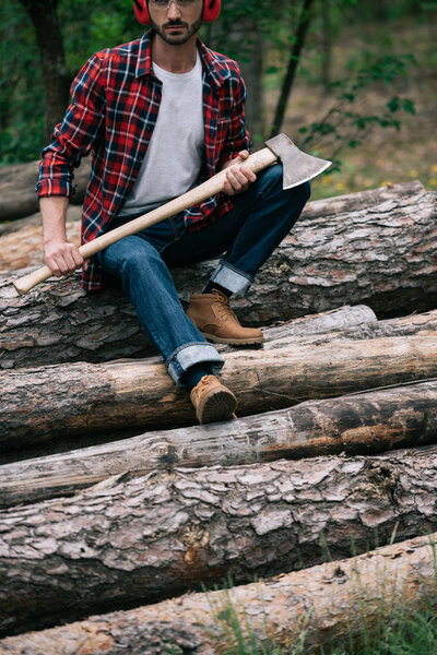 patial view of lumberer sitting on logs in forest and holding ax