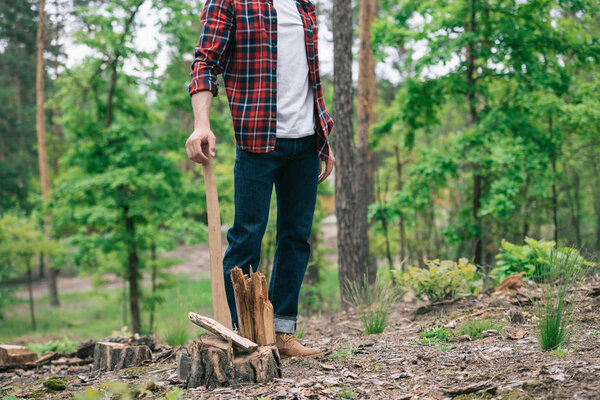partial view of lumberjack in plaid shirt and denim jeans standing with ax in forest