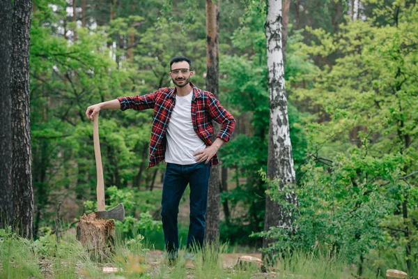 smiling lumberjack in plaid shirt and denim jeans holding ax while standing with hand on hip and looking at camera