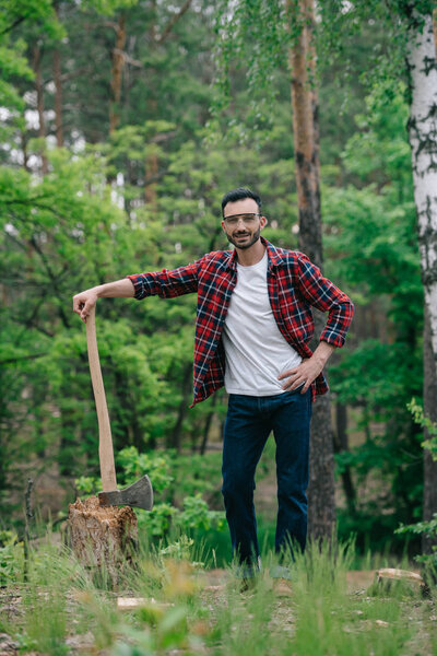 cheerful lumberjack in plaid shirt and denim jeans holding ax while standing with hand on hip and smiling at camera